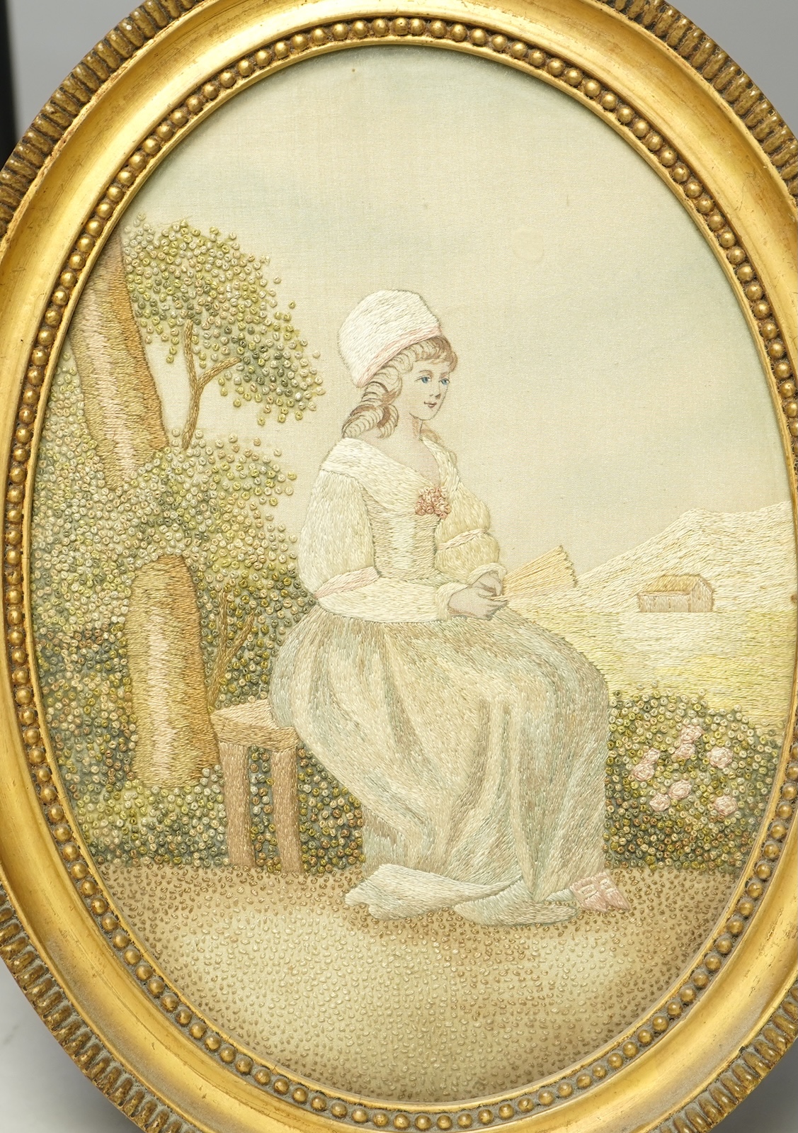 A pair of early 20th century small gilt oval embroideries, of a girl gathering fruit from a tree and a seated lady in a landscape, worked in stem stitch and decorative knotting. 29cm long including gilt frames. Condition
